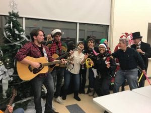 Officers in festive holiday gear join a guitarist and sing next to a Christmas tree in a community center space.