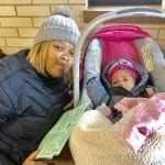 A mother in winter gear crouches next to her newborn child in a crib