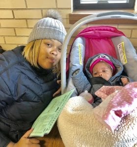 A mother in winter gear crouches next to her newborn child in a crib