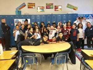 An officer bends over a table to pose with elementary school children, who are smiling and making faces.