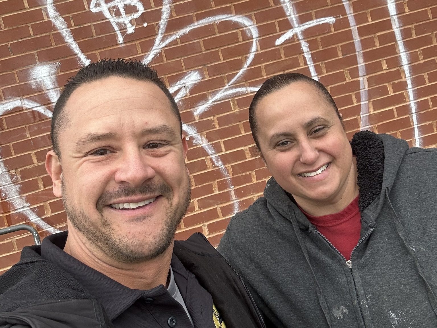 Commander Maffo-Judd and Sandra smiling in front of a brick wall partially covered in graffiti, following Sandra’s volunteer cleanup efforts