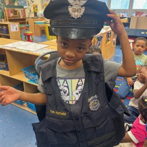 Child tips a patrol cap while wearing a vest with “The Future” patch.