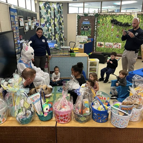 Students gather around a seated Easter Bunny and community officers in a colorful classroom setting.
