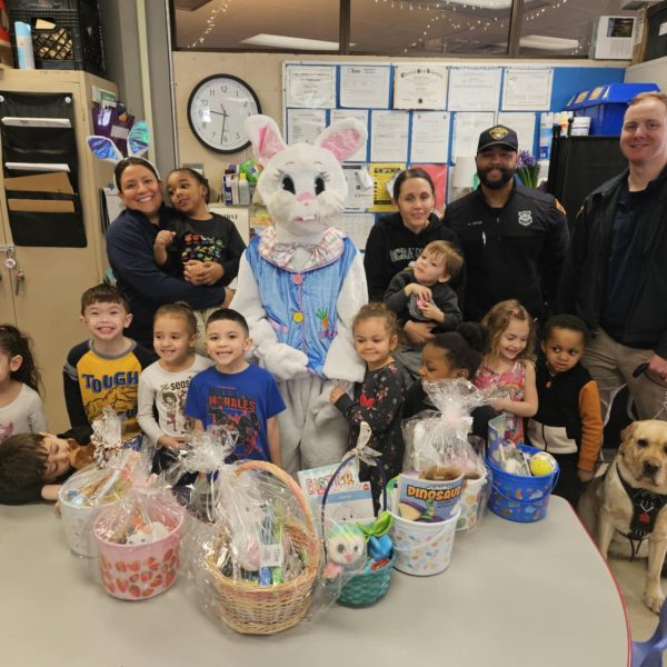 Children and staff smile with the Easter Bunny and DOP officers, surrounded by gift baskets and a therapy dog.