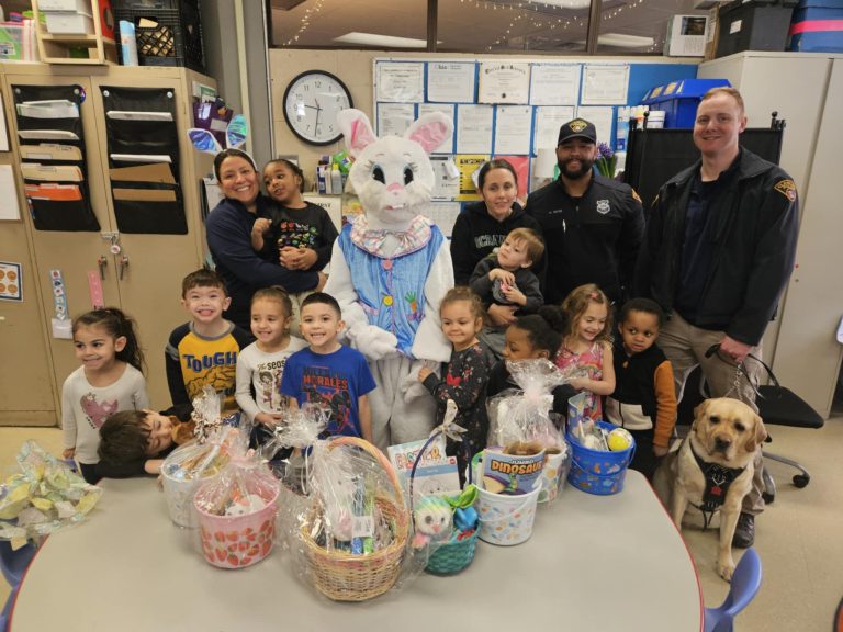 Children and staff smile with the Easter Bunny and DOP officers, surrounded by gift baskets and a therapy dog.