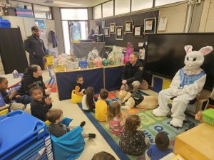 Young students seated on a reading rug listen as officers visit with Easter baskets and the Bunny.