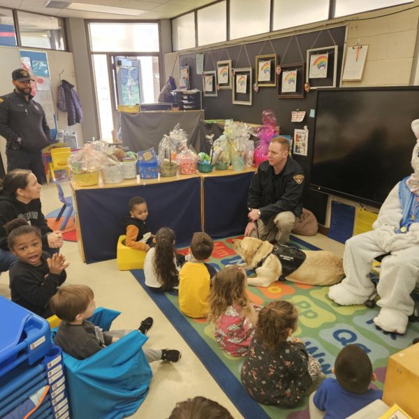 Young students seated on a reading rug listen as officers visit with Easter baskets and the Bunny.