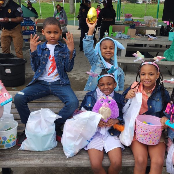 Group of children sit on a bench showing off Easter toys and baskets, with officers and families visible in the background.
