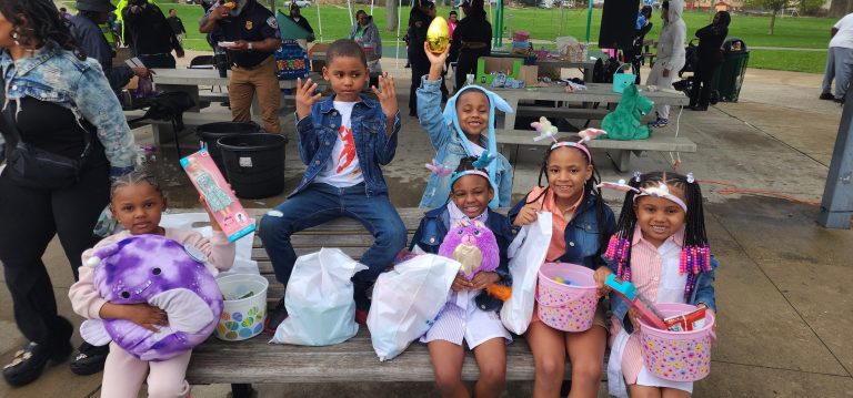 Group of children sit on a bench showing off Easter toys and baskets, with officers and families visible in the background.