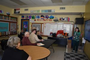 A student at the front of a colorful elementary classroom presents while officers and other adults observe attentively.