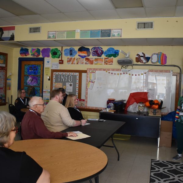A student at the front of a colorful elementary classroom presents while officers and other adults observe attentively.
