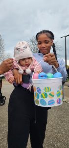 Woman holds a baby wearing a bunny hat, both looking into a pastel egg-filled basket.