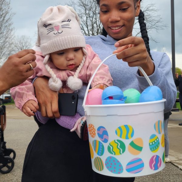 Woman holds a baby wearing a bunny hat, both looking into a pastel egg-filled basket.