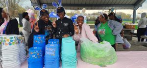 Volunteers in bunny ears smile and flash peace signs behind stacks of colorful Easter baskets at a park pavilion.