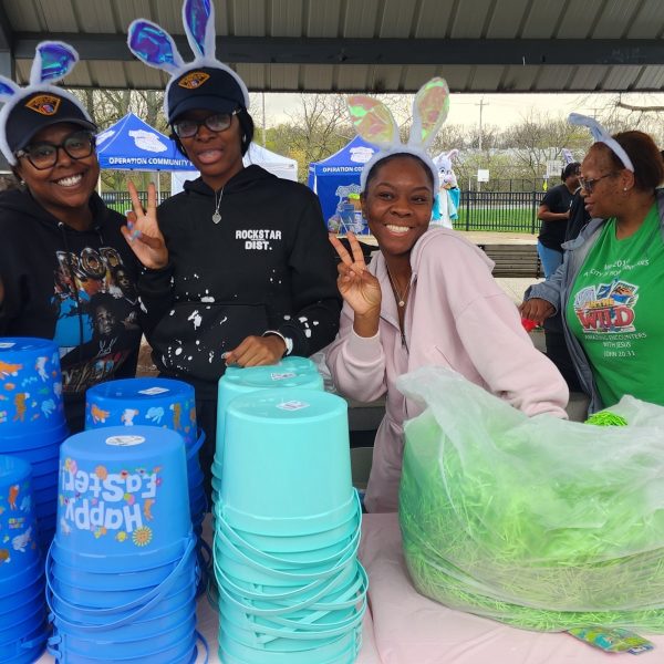 Volunteers in bunny ears smile and flash peace signs behind stacks of colorful Easter baskets at a park pavilion.
