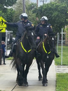 Two Mounted Unit officers ride side-by-side down the sidewalk as students watch.