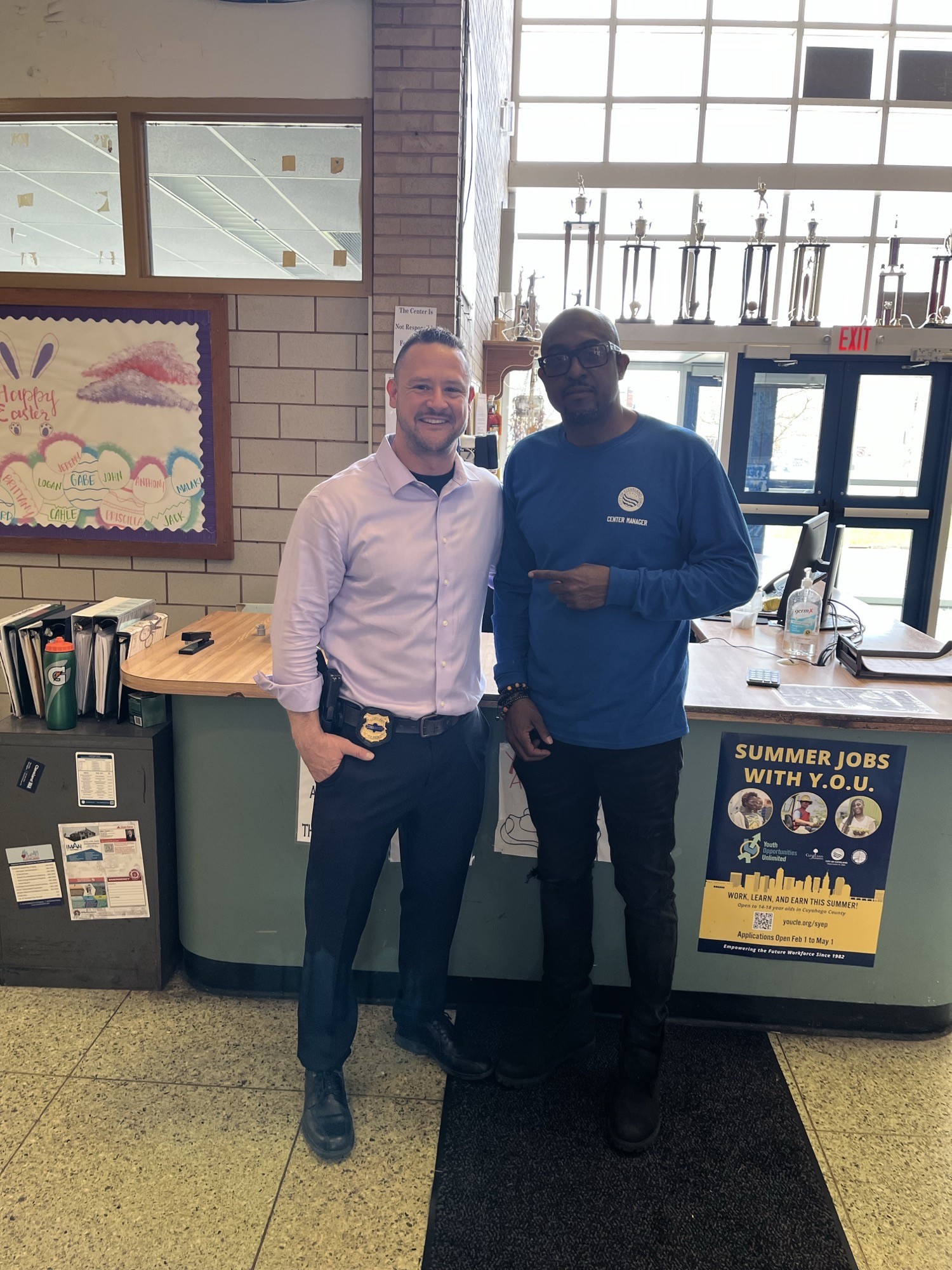 Commander Maffo-Judd stands with Estabrook Rec Center Director Jack Nichols near the front desk, with trophies and a youth job flyer in the background.