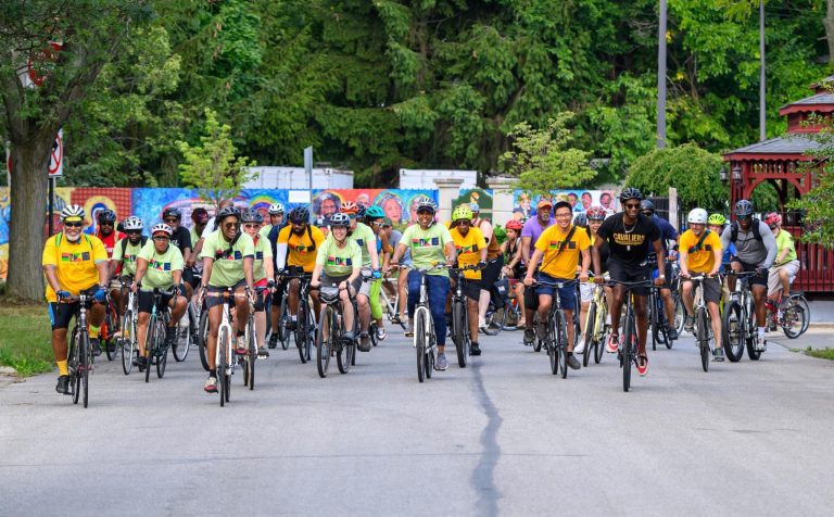A large group of Bike Your Neighborhood riders in matching shirts and helmets rolls out together from a park, with a colorful mural and trees in the background