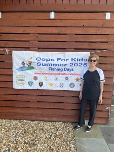 A volunteer stands beside a “Cops for Kids Summer 2025 Fishing Days” banner displayed on a wooden wall.
