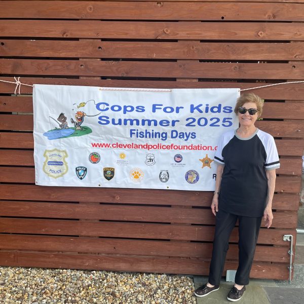 A volunteer stands beside a “Cops for Kids Summer 2025 Fishing Days” banner displayed on a wooden wall.