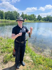 A smiling officer holds up a fishing pole and small fish by the edge of the lake on a sunny day.