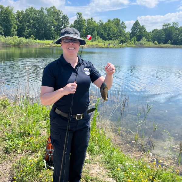 A smiling officer holds up a fishing pole and small fish by the edge of the lake on a sunny day.