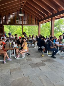 Kids and adults seated at picnic tables enjoy lunch together under the park pavilion.