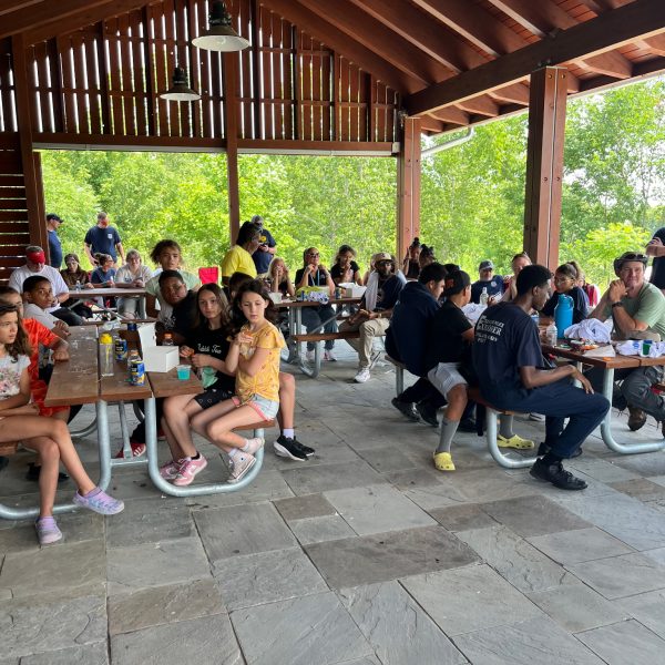 Kids and adults seated at picnic tables enjoy lunch together under the park pavilion.