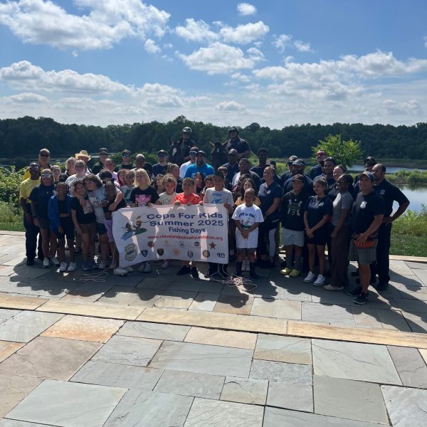 A large group of officers, kids, and community members pose together with the event banner near the water.