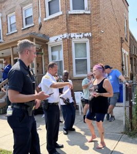 Sergeant Thomas and Commander Maffo-Judd speak with a barefoot woman with purple hair outside a brick apartment building.