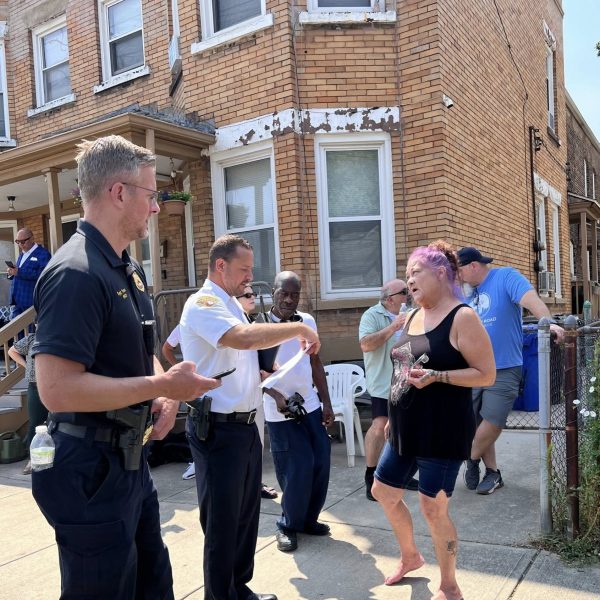 Sergeant Thomas and Commander Maffo-Judd speak with a barefoot woman with purple hair outside a brick apartment building.