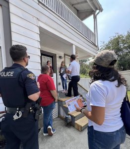 Officers and community partners visit a resident on her porch during the safety walk, with one woman holding a Trent Park flyer.