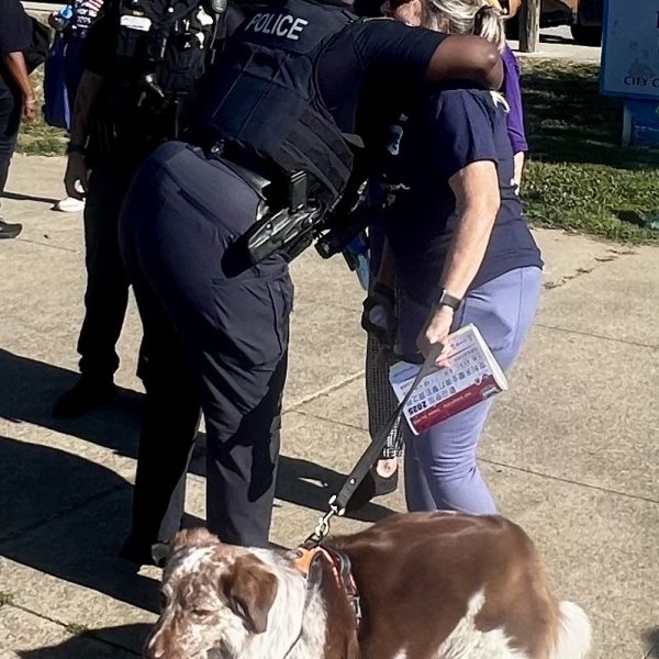 Officer hugs a senior woman with a dog on a leash while other participants and officers look on nearby.