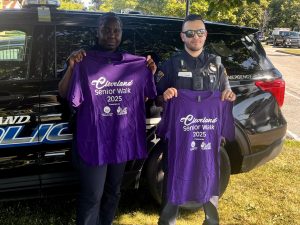 Two Cleveland police officers pose holding up 2025 Cleveland Senior Walk t-shirts in front of a police SUV.