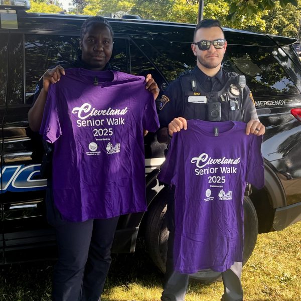 Two Cleveland police officers pose holding up 2025 Cleveland Senior Walk t-shirts in front of a police SUV.