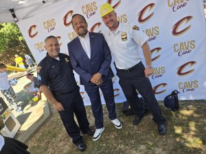 Three men pose in front of a Cavs Care backdrop, including two Cleveland officers and a suited event guest wearing a Cavaliers hat.