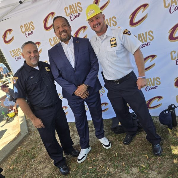 Three men pose in front of a Cavs Care backdrop, including two Cleveland officers and a suited event guest wearing a Cavaliers hat.
