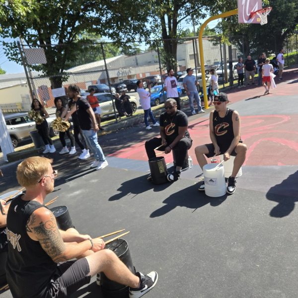 Members of a Cavs drumline perform on plastic buckets while cheerleaders and event attendees gather along the court perimeter.