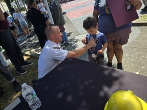 A Cleveland police officer kneels to talk with a young child wearing Cavs-themed medallions and holding sunglasses, while the child's guardian stands nearby.
