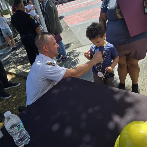 A Cleveland police officer kneels to talk with a young child wearing Cavs-themed medallions and holding sunglasses, while the child's guardian stands nearby.