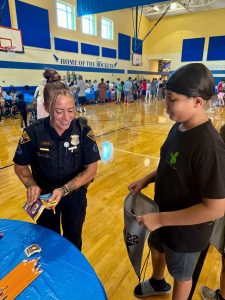 An officer smiles while giving a pack of crayons to a student holding a drawstring bag during the school supply event at RG Jones School.