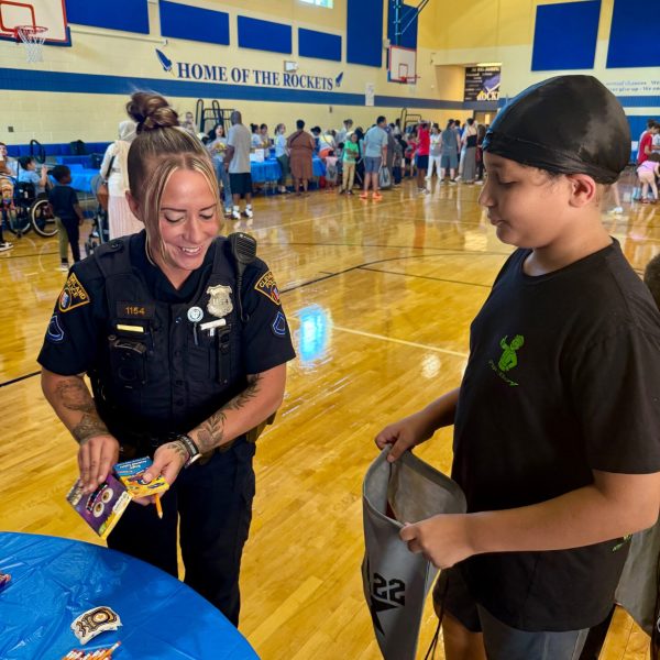 An officer smiles while giving a pack of crayons to a student holding a drawstring bag during the school supply event at RG Jones School.