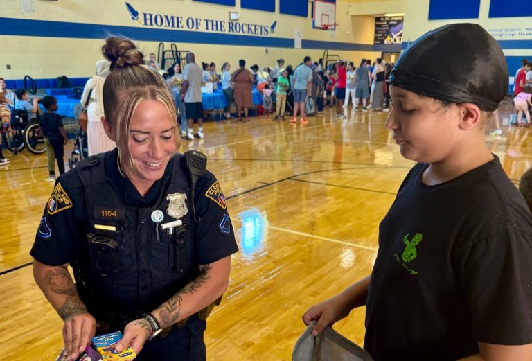 An officer smiles while giving a pack of crayons to a student holding a drawstring bag during the school supply event at RG Jones School.