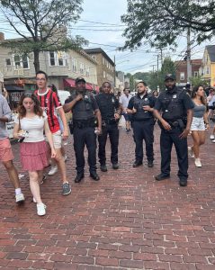 Four Cleveland Police officers stand on a brick-paved street in Little Italy during the Feast of the Assumption, surrounded by festival crowds.
