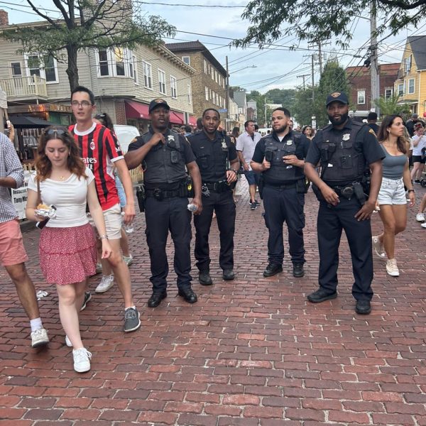 Four Cleveland Police officers stand on a brick-paved street in Little Italy during the Feast of the Assumption, surrounded by festival crowds.
