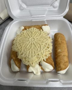 A takeout container holds fresh Italian pastries including cannoli filled with cream and a decorative pizzelle cookie.