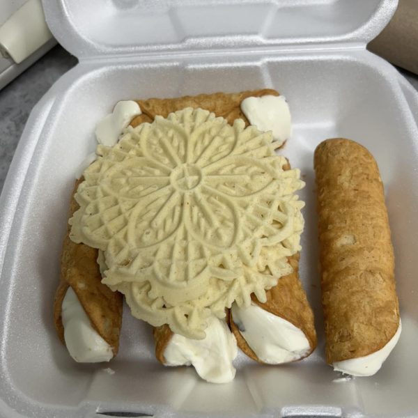 A takeout container holds fresh Italian pastries including cannoli filled with cream and a decorative pizzelle cookie.