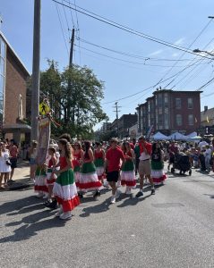 Parade participants dressed in red, white, and green skirts and shirts march down Mayfield Road as crowds look on during the Feast of the Assumption.
