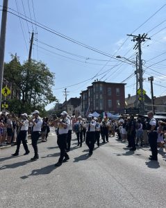 A marching band in white shirts and dark pants plays instruments as they proceed down Mayfield Road during the Feast of the Assumption parade.
