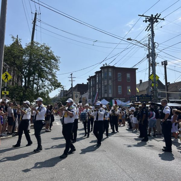 A marching band in white shirts and dark pants plays instruments as they proceed down Mayfield Road during the Feast of the Assumption parade.